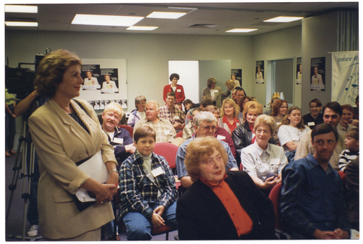 COL photo of a room full of people sitting on rows of chairs. One woman is standing in the foreground, to the left of the frame, and a few other people are standing at the back of the room. Behind the woman in the foreground, there is a camera operaor behind a television camera on a stand (partially obscured). On the walls around the room, there are several posters depicting the image of the woman standing in the foreground.