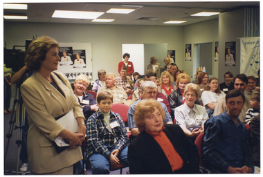 COL photo of a room full of people sitting on rows of chairs. One woman is standing in the foreground, to the left of the frame, and a few other people are standing at the back of the room. Behind the woman in the foreground, there is a camera operaor behind a television camera on a stand (partially obscured). On the walls around the room, there are several posters depicting the image of the woman standing in the foreground.