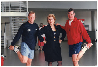 COL photo of three people posing for a photo. A woman (in the centre) is wearing a suit jacket. She has her hands on her hips and is turning slightly to one side. Another woman (wearing a track suit jacket) and a man (wearing a sweatshit) are standing on either side of her. They each have both of their hands placed on one of her shoulders. The trio are standing in the large lobby of a gymnasium or stadium. There are two other people in the background.