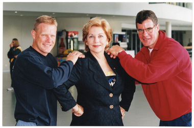COL photo of three people posing for a photo. A woman (in the centre) is wearing a suit jacket and is turning slightly to one side. Two men (standing on either side of her) are wearing sweatshirts. They each have both of their hands placed on one of her shoulders. The trio is standing in the large lobby of a gymnasium or stadium. In the background, there is another person wearing a green and gold track suit, and some vending machines along the wall.