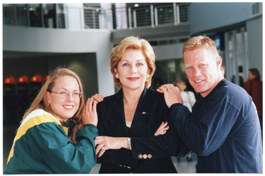 COL photo of three people posing for a photo. A woman (in the centre) is wearing a suit jacket. She has her arms crossed across her chest. Another woman (wearing a track suit jacket) and a man (wearing a sweatshit) are standing on either side of her. They each have both of their hands placed on one of her shoulders. The trio are standing in the large lobby of a gymnasium or stadium. There are two other people in the background.