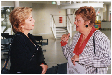 COL photo of two women standing close together, chatting. They are in the large lobby of a gymnasium or stadium.