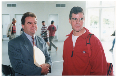 COL photo of two men standing close together. They are in the large lobby of a gymnasium or stadium. In the background, there are a few people walking through the lobby.