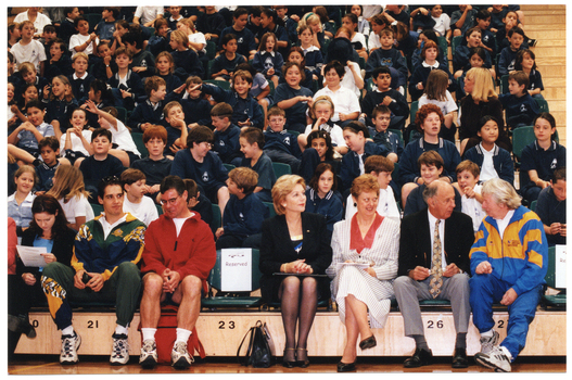 COL photo of several adults seated in the front row of multiple rows of tiered seating in a gymnasium or stadium. Seated behind them are several rows of school children in school uniform.