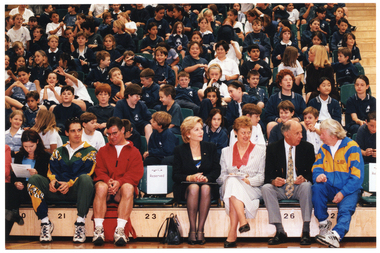 COL photo of several adults seated in the front row of multiple rows of tiered seating in a gymnasium or stadium. Seated behind them are several rows of school children in school uniform.