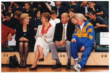 COL medium close up photo of several adults seated in the front row of multiple rows of tiered seating in a gymnasium or stadium. Seated behind them are several rows of school children in school uniform.