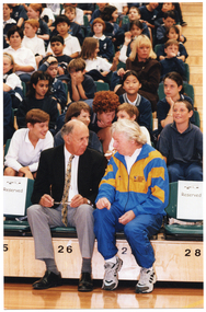 COL medium close up photo of two men seated in the front row of multiple rows of tiered seating in a gymnasium or stadium. Seated behind them are several rows of school children in school uniform.
