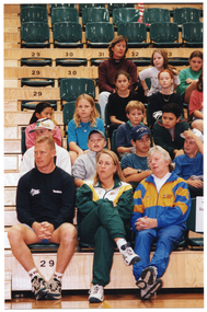 COL medium close up photo of three adults seated in the front row of multiple rows of tiered seating in a gymnasium or stadium. Seated behind them are several rows of school children in school uniform.