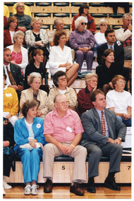 COL medium close up photo of three adults seated in the front row of multiple rows of tiered seating in a gymnasium or stadium. Seated behind them are several rows of people. Some of them are wearing name tags or badges.