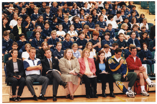 COL wide frame photo of several adults seated in the front row of multiple rows of tiered seating in a gymnasium or stadium, Seated behind them are several rows of school children in school uniform.