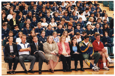 COL wide frame photo of several adults seated in the front row of multiple rows of tiered seating in a gymnasium or stadium, Seated behind them are several rows of school children in school uniform.