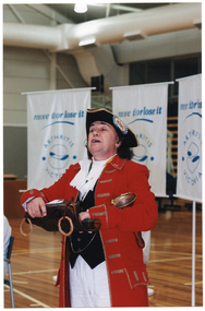 COL medium close up photo of a man dressed in a traditional costume, standing in a gymnasium or stadium. His costume consists of a white shirt with a frilled lace neck, a black vest, a red jacket with gold brocade trimming, white trousers, and a black hat with gold brocade trim and a large rosette featuring a coat of arms design in the centre. He is unrolling a leather scroll and has a brass bell tucked between his torso and the crook of one arm. Behind him, there are three freestanding vertical banners in white with the text, 'Move It Or Lose It', in blue, followed by the text and logo for 'Arthritis Victoria'.