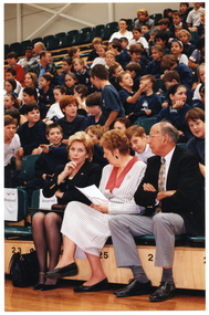 COL photo of three people seated in the front row of multiple rows of tiered seating in a gymnasium or stadium. There are several rows of school children in school uniform seated behind them.