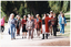 COL photo of several people walking along a path in a large park. One of them is a man dressed in a traditional costume, while everyone else is dressed in contemporary clothes. The man in costume is holding a large brass bell and a leather scroll.