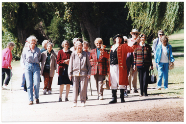 COL photo of several people walking along a path in a large park. One of them is a man dressed in a traditional costume, while everyone else is dressed in contemporary clothes. The man in costume is holding a large brass bell and a leather scroll.
