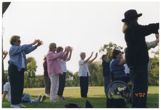 COL side-view photo of a group of people doing some stretching exercises in a park. One person is a wheelchair user and another person is sitting on the grass. Most people are holding their arms outstretched in front on them.