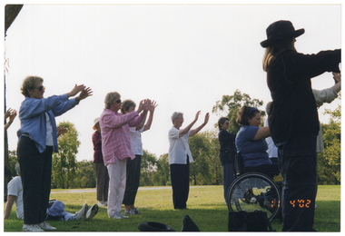 COL side-view photo of a group of people doing some stretching exercises in a park. One person is a wheelchair user and another person is sitting on the grass. Most people are holding their arms outstretched in front on them.