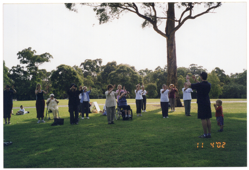 COL wide frame photo of a group of people, led by an instructor, doing some stretching exercises in a park. They have their hands outstretched in front of them, at shoulder or chest height. One person is a wheelchair user, while another is sitting on a walking frame. There are a few young children amongst the group of adults.