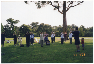 COL wide frame photo of a group of people, led by an instructor, doing some stretching exercises in a park. They have their hands outstretched in front of them, at shoulder or chest height. One person is a wheelchair user, while another is sitting on a walking frame. There are a few young children amongst the group of adults.