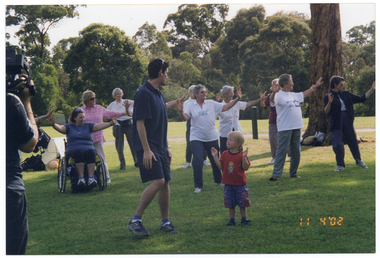 COL photo of a group of people, led by an instructor, doing some stretching exercises in a park. One of them is a wheelchair user. They have their arms outstretched wide in front of them. Most of those standing are leaning to one side with their weight on one leg. There is a young child in the foreground. To the left of the frame (partially obscured), there is a television camera operator with a camera on his shoulder.