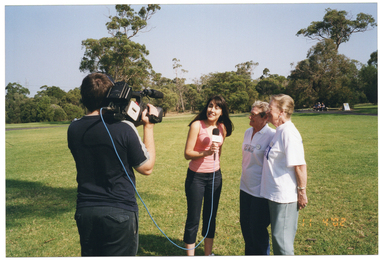 COL photo of a small group of people in a park. One is a camera operator who has a television camera sitting on top of his shoulder. He is filming three women who are standing opposite him. One is a young woman. She is facing the camera while holding a microphone on a lead, which is attached to the camera. Two middle-aged women are standing next to her, facing her.