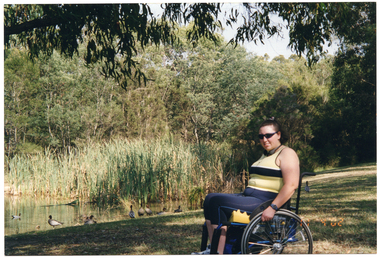 COL photo of a woman posing for a photo beneath a large shady tree by a pond or lake. She is a wheelchair user. There are some ducks at the edge of the lake.