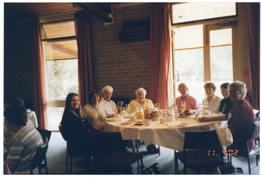 COL photo of a group of people sitting at a large round table. There is food and drinks on the table. Another group of people (partially obscured) are sitting at another table to the left of the frame. There is a view of the park through the windows.