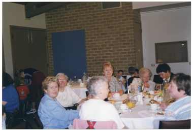 COL photo of a group of people sitting at a large round table. There is food and drinks on the table. Another group of people (partially obscured) are sitting at another table to the left of the frame. In the background, there is a woman attending to a young child near the wall.