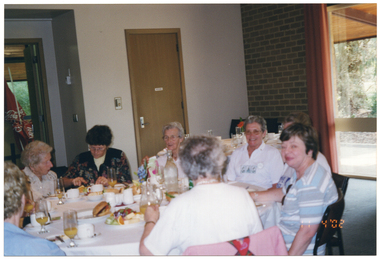 COL photo of a group of people sitting at a large round table. There is food and drinks on the table. In the background, there are some red table umbrellas with the "Coca-Cola" logo leaning against the wall in an adjoining room. There is a view of some trees through a large window.
