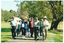 COL photo of a group of people walking along a concrete path at a park. They are walking directly towards the camera and smiling. One person at the centre front of the group is holding a sheet of paper in one hand and waving with her other hand.
