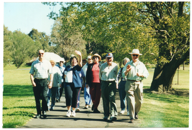 COL photo of a group of people walking along a concrete path at a park. They are walking directly towards the camera and smiling. One person at the centre front of the group is holding a sheet of paper in one hand and waving with her other hand.