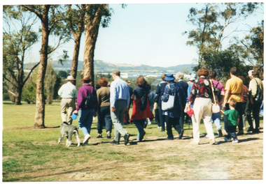COL photo of a group of people walking along a path at a park. One person has a dog on a leash. Another person is holding the hand of a small child. There are some hills in the distance, beyond the park. The photo has been taken from behind the walking group.