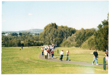 COL wide frame photo of a group of people walking around the bend of a path at a park. There are some hills in the distance, beyond the park. The photo has been taken from a distance.