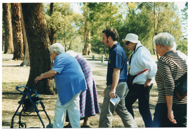 COL close up photo, taken from the side, of a group of people walking along a path at a park. One person is using a walking frame.