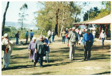 COL photo of a group of people gathered on the grassy area near some shelters or the veranda of a building. Some people are walking, while others are standing. A few people are using walking sticks, and one person (partially obscured) is using a walking frame. In the background, there is a person with a dog on a leash.