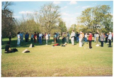 COL photo of a group of people gathered on the grass at a park. Most people are standing, facing one person standing in front of the group. One person is sitting down on the grass, slightly further away, also facing the group. There are several large trees in the background. The photo has been taken from a slight angle, with most of the group in three-quarter profile.