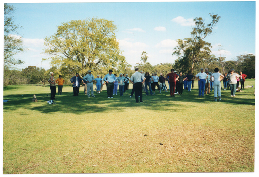 COL wide frame photo of a group of people standing on the grass at a park. They are facing two people, who are also standing and, in turn, facing the group. The photo has been taken from behind the two people facing the group.