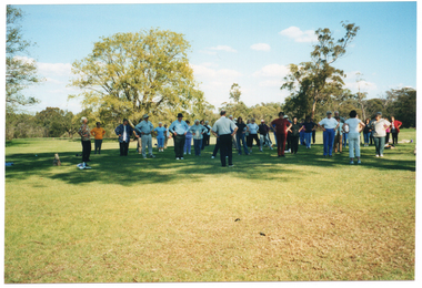 COL wide frame photo of a group of people standing on the grass at a park. They are facing two people, who are also standing and, in turn, facing the group. The photo has been taken from behind the two people facing the group.