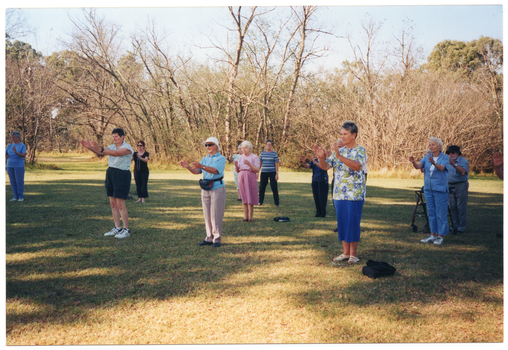 COL photo of a group of people standing in a large park, doing some exercises. They are standing slightly apart on a grassy area, with their hands up in front of their chests. One participant is standing behind his walking frame. There are many trees in the background.