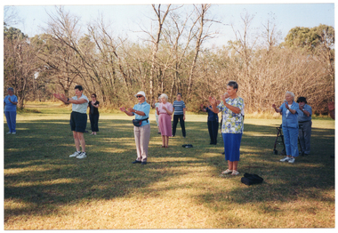 COL photo of a group of people standing in a large park, doing some exercises. They are standing slightly apart on a grassy area, with their hands up in front of their chests. One participant is standing behind his walking frame. There are many trees in the background.