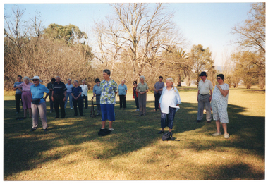 COL photo of a group of people standing in a large park, doing some exercises. They are standing slightly apart on a grassy area, with many trees in the background. One participant is standing behind his walking frame.