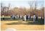 COL photo of a group of people standing on a grassy area in a large park, doing some exercises. One woman is standing apart from the group, facing them. Everyone is standing slightly apart , with their hands up in front of their chests. One participant is standing behind his walking frame. There are many trees in the background.