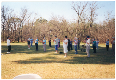 COL photo of a group of people standing on a grassy area in a large park, doing some exercises. One woman is standing apart from the group, facing them. Everyone is standing slightly apart , with their hands up in front of their chests. One participant is standing behind his walking frame. There are many trees in the background.