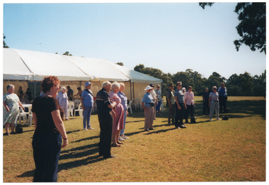 COL photo of a group of people standing on a grassy area in front of a large marquee. They are standing with their arms by their sides. In the background, there are some other people standing inside the marquee, in which there are several white plastic tables and chairs set up.