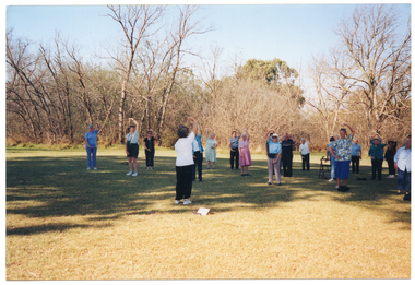 COL photo of a group of people standing on a grassy area in a large park. In this photo, a woman is standing apart from the group, facing them.  One participant is standing behind his walking frame. Everyone is holding one curved arm up above their heads, with their other curved arm down in front of their pelvises. There are many trees in the background.
