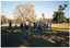 COL photo of a group of people standing on a grassy area in a large park. They are standing slightly apart, and lunging slightly forward on one leg while holding their arms out in front of them. One participant is standing behind his walking frame.