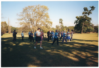 COL photo of a group of people standing on a grassy area in a large park. They are standing slightly apart, and lunging slightly forward on one leg while holding their arms out in front of them. One participant is standing behind his walking frame.