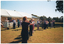 COL photo of a group of people standing on a grassy area in front of a large white marquee. They are holding their curved arms outstretched, with hands almost touching. There are other people inside the marquee, with white plastic tables and chairs around them. The photo has been taken at a slight angle from the side. One woman in the foreground has her back to the camera.