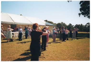 COL photo of a group of people standing on a grassy area in front of a large white marquee. They are holding their curved arms outstretched, with hands almost touching. There are other people inside the marquee, with white plastic tables and chairs around them. The photo has been taken at a slight angle from the side. One woman in the foreground has her back to the camera.