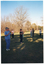 COL photo of a group of people standing on a grassy area in a large park. They are standing slightly apart, with their hands up in front of their chests. There are many trees in the background.